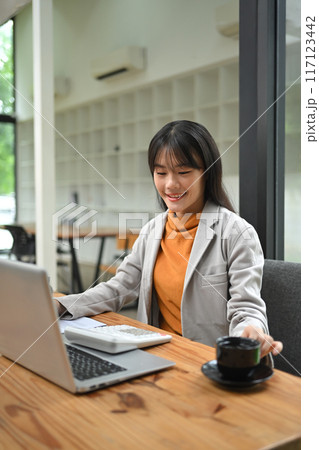 Smiling young businesswoman working with laptop and financial reports at modern office 117123442