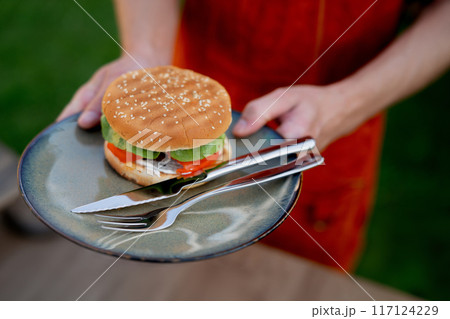 Close up of man holding plate with homemade grilled hamburger and cutlery. Summer outdoor barbecue. 117124229