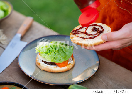 Close up of man assembling burgers with toppings, putting barbecue sauce on toasted bun. Summer outdoor barbecue. Close up of man assembling burgers with toppings, putting barbecue sauce on toasted bun. Summer outdoor barbecue. 117124233