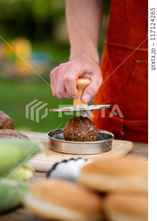 Man in apron preparing meat for hamburgers, standing outdoors. Using burger patty press to shape groud meat. 117124265