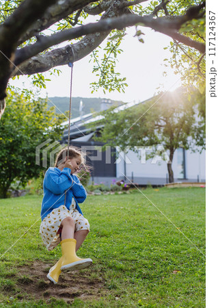 A young girl swinging on a swing in the garden. The schoolgirl spending summer holiday with her grandparents in countryside. 117124367