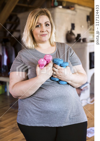 Overweight woman exercising at home, holding blue and pink dumbbells. Workout in living room. Overweight woman exercising at home, holding blue and pink dumbbells. Workout in living room. 117124433