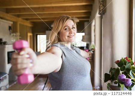 Overweight woman exercising at home, using dumbbells. Workout in living room. Overweight woman exercising at home, using dumbbells. Workout in living room. 117124446