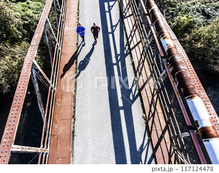 Aerial view of a two runners running across bridge over the river. Morning running training for male friends. Aerial view of a two runners running across bridge over the river. Morning running training for male friends. 117124478