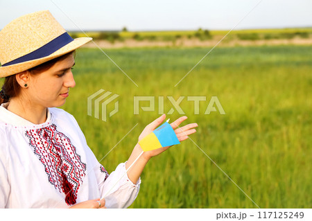 Portrait of a young Ukrainian woman against a meadow nature background. She is smiling and showing a gesture of love, while holding the Ukrainian flag. Copy space 117125249