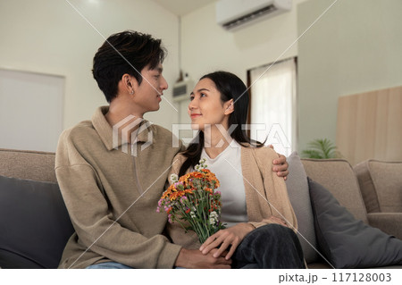 Young Couple Sitting on Sofa with Flowers, Sharing a Loving Moment in a Modern Living Room 117128003
