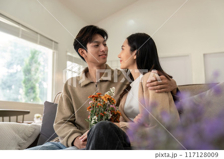 Romantic Couple Sitting on Sofa with Flowers in Cozy Living Room, Sharing a Tender Moment 117128009