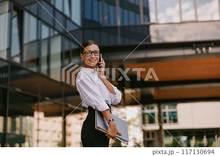 In an urban setting, a businesswoman is on a professional call, displaying her elegance In an urban setting, a businesswoman is on a professional call, displaying her elegance 117130694