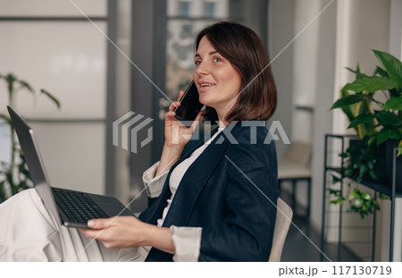 Professional businesswoman at work, smiling with phone and laptop, showing confidence and success Professional businesswoman at work, smiling with phone and laptop, showing confidence and success 117130719