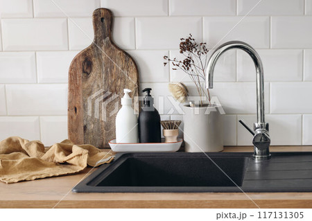 Scandinavian kitchen interior. Wooden countertop, chopping board. Soap, detergent dispenser glass bottles mockup. Dish brush, black sink. Eco dish washing concept. White wall, metro tiles background Scandinavian kitchen interior. Wooden countertop, chopping board. Soap, detergent dispenser glass bottles mockup. Dish brush, black sink. Eco dish washing concept. White wall, metro tiles background 117131305