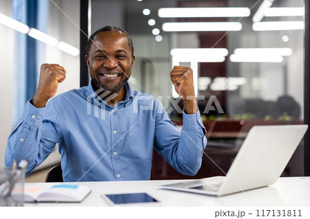 Happy african american office worker celebrating success at workplace. Mature businessman smiling with clenched fists in modern office. Achievement, happiness, and professional success concept. Happy african american office worker celebrating success at workplace. Mature businessman smiling with clenched fists in modern office. Achievement, happiness, and professional success concept. 117131811