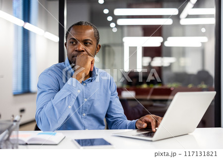 Pensive office worker in blue shirt thinking at desk with open laptop and documents. Modern office environment with bright lights and large windows. Concept of contemplation, problem-solving 117131821