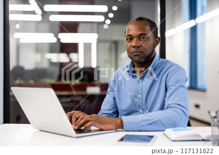 African American businessman sitting at desk with laptop inside modern office. Professional workplace setting with businessman focusing on work. Office setting promotes productivity professionalism. African American businessman sitting at desk with laptop inside modern office. Professional workplace setting with businessman focusing on work. Office setting promotes productivity professionalism. 117131822