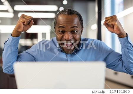 Happy businessman celebrating success at work on laptop. Man with raised fists showing joy and excitement while looking at screen. Concept of achievement, victory, and career success. Happy businessman celebrating success at work on laptop. Man with raised fists showing joy and excitement while looking at screen. Concept of achievement, victory, and career success. 117131860
