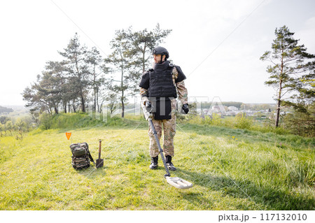 A man in a military uniform and bulletproof vest works in the forest with a metal detector. A minesweeper performs work on demining the territory 117132010