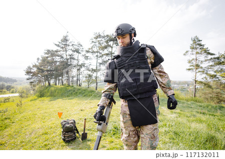 A man in a military uniform and bulletproof vest works in the forest with a metal detector. A minesweeper performs work on demining the territory 117132011