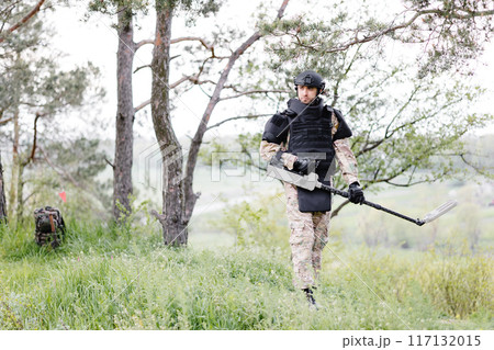 A man in a military uniform and bulletproof vest works in the forest with a metal detector. A minesweeper performs work on demining the territory 117132015