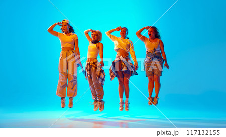 Dancers leap into air, in playful salute pose against vibrant blue background in neon light, shows their synchronized movements and casual streetwear style. 117132155