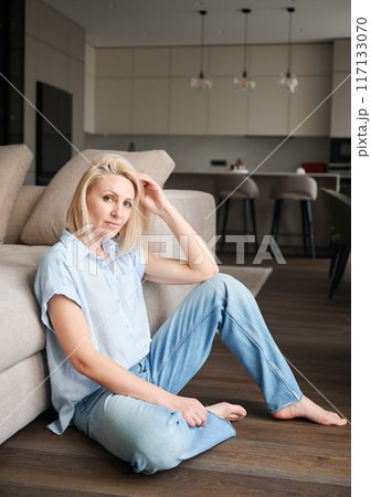 Gorgeous woman sitting next to couch in her apartment. Pretty female in casual clothes looking straight on camera. Gorgeous woman sitting next to couch in her apartment. Pretty female in casual clothes looking straight on camera. 117133070