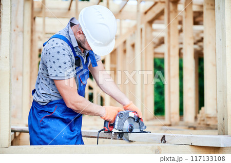 Carpenter using circular saw for cutting wooden plank. Man worker building wooden frame house. The idea behind modern ecological construction. 117133108