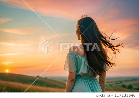 夕暮れの草原で空を見つめている青いドレスの女性の後姿 117133640