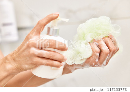 female hands pouring liquid moisturizing soap shower gel from white cosmetic bottle on bath puff in bathroom 117133778