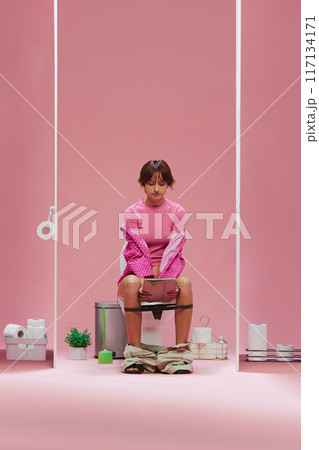 Young girl in pink clothes, with short hair sitting on toilet and looking on tablet against pink background 117134171
