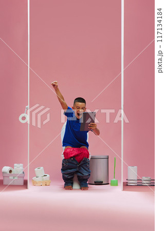Winning game. Young boy, child sitting in toilet and watching soccer game online on tablet, emotionally supporting favorite team against pink background 117134184