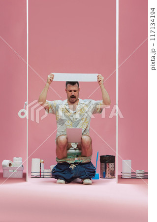 Man sitting on toilet, raising toilet paper upwards and watching soccer game event online via tablet against pink background 117134194