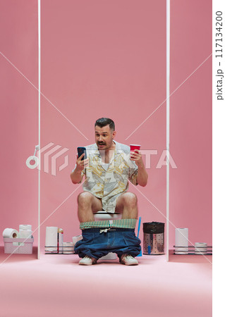 Excited man sitting in toilet with cup of drink and mobile phone, watching online soccer game translation with excitement against pink background 117134200
