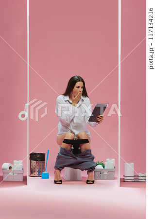 Young woman, office worker sitting in toilet with tablet and watching soccer game translation with shocked face against pink background Young woman, office worker sitting in toilet with tablet and watching soccer game translation with shocked face against pink background 117134216