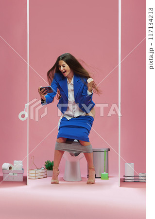 Young woman in blue suit standing in toilet room and emotionally watching online soccer game via mobile phone against pink background Young woman in blue suit standing in toilet room and emotionally watching online soccer game via mobile phone against pink background 117134218