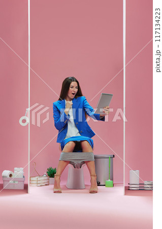 Successful game. Young emotional woman in formal wear sitting in toilet and watching live game via tablet against pink background Successful game. Young emotional woman in formal wear sitting in toilet and watching live game via tablet against pink background 117134223