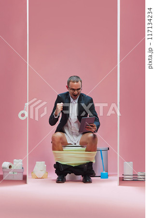 Businessman in suit sitting on toilet with pants down and emotionally watching online soccer game translation via tablet against pink background Businessman in suit sitting on toilet with pants down and emotionally watching online soccer game translation via tablet against pink background 117134234