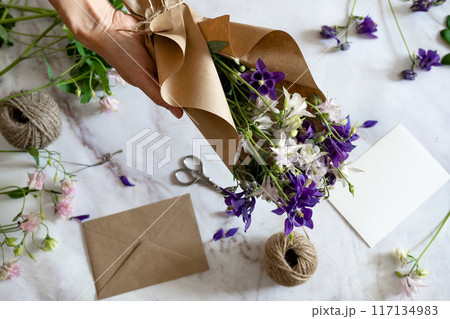 Closeup of hands of young woman florist creating invitation, put in envelope blossom flowers, top view. Macro. Isolated on white background. With free space for your text 117134983