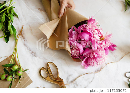 A female florist makes a bouquet of peony flowers wrapped in kraft paper on her desktop. View from above. High quality photos 117135072