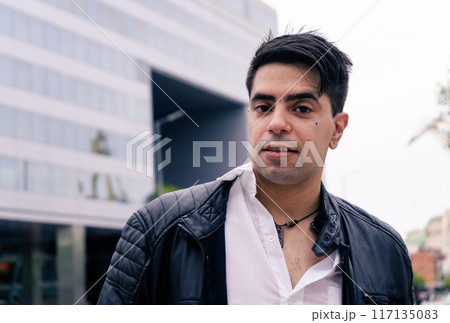 Attractive Latino man in a black jacket and white shirt with a modern and important building behind. Attractive Latino man in a black jacket and white shirt with a modern and important building behind. 117135083