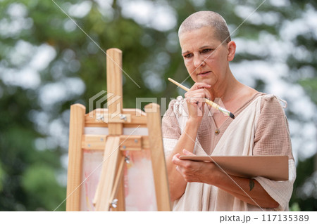 Thoughtful Artist Contemplating Her Work While Holding a Paintbrush and Palette Outdoors 117135389