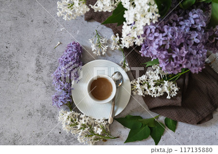 The postcard is beautiful. A fancy purple coffee mug, an old book, a straw hat and a bouquet of purple lilac. Beautiful still life. Spring time. The concept of Good morning. 117135880