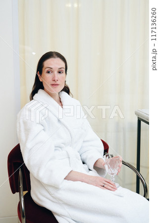 Relaxing Woman in a White Bathrobe Sitting While Holding a Glass in a Bright Indoor Space Relaxing Woman in a White Bathrobe Sitting While Holding a Glass in a Bright Indoor Space 117136260