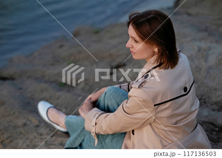 A beautiful girl sits on the seashore and looks into the distance. Her hand draws a drawing in the sand 117136503