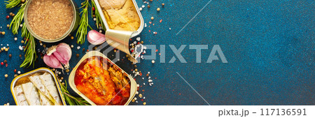 Different open tin cans with canned fish like salmon, mackerel, sprat, sardine, fish pate, among spices and herbs on a blue background, banner 117136591