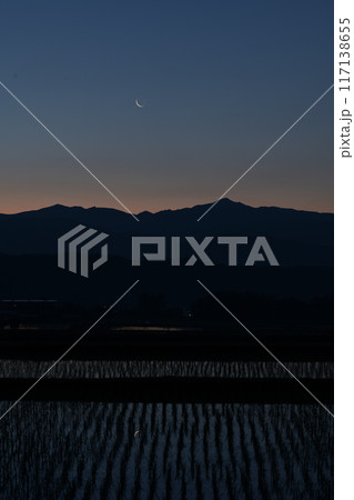 北杜市白州町から見る奥秩父山塊上の月齢28.0の細い有明の月を映す水田の苗間 117138655