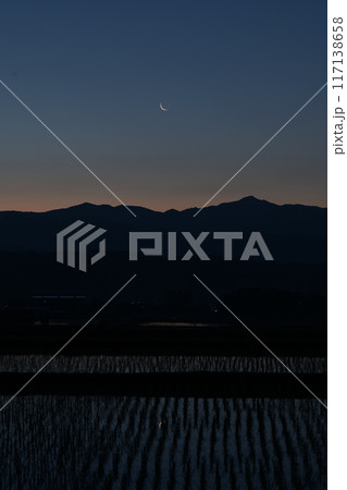 北杜市白州町から見る奥秩父山塊上の月齢28.0の細い有明の月を映す水田の苗間 117138658