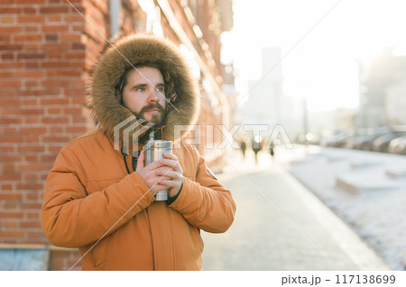 Close-up portrait handsome bearded millennial man in winter clothes and with thermos snow in city copy space. Cold season and hot beverage in winter time 117138699
