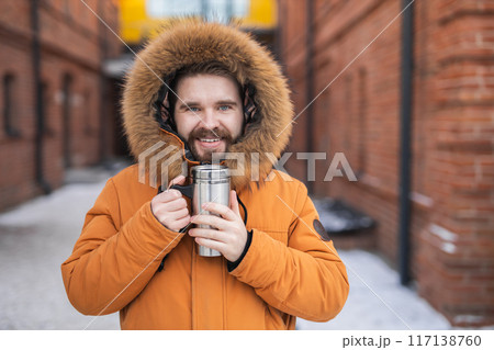 Close-up portrait handsome bearded millennial man in winter clothes and with thermos snow outdoor. Cold season and hot beverage in winter time Close-up portrait handsome bearded millennial man in winter clothes and with thermos snow outdoor. Cold season and hot beverage in winter time 117138760