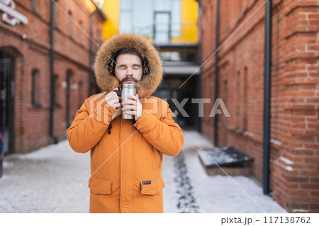 Close-up portrait handsome bearded millennial man in winter clothes and with thermos snow in city. Cold season and hot beverage in winter time 117138762