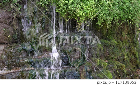 A serene waterfall flows gently down rocky terrain surrounded by greenery A serene waterfall flows gently down rocky terrain surrounded by greenery 117139052