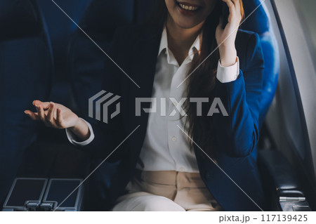 A young Asian female airplane passenger sits by the window during the flight, holding her boarding pass, with her carry-on luggage stored overhead, wearing her seatbelt, and enjoying the journey. 117139425
