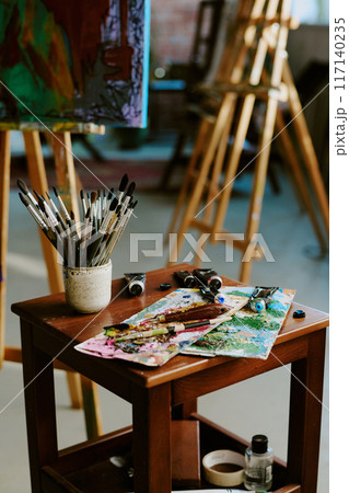 Vertical shot of set of paintbrushes, paint tubes, palettes on small wooden table in modern art studio or school Vertical shot of set of paintbrushes, paint tubes, palettes on small wooden table in modern art studio or school 117140235
